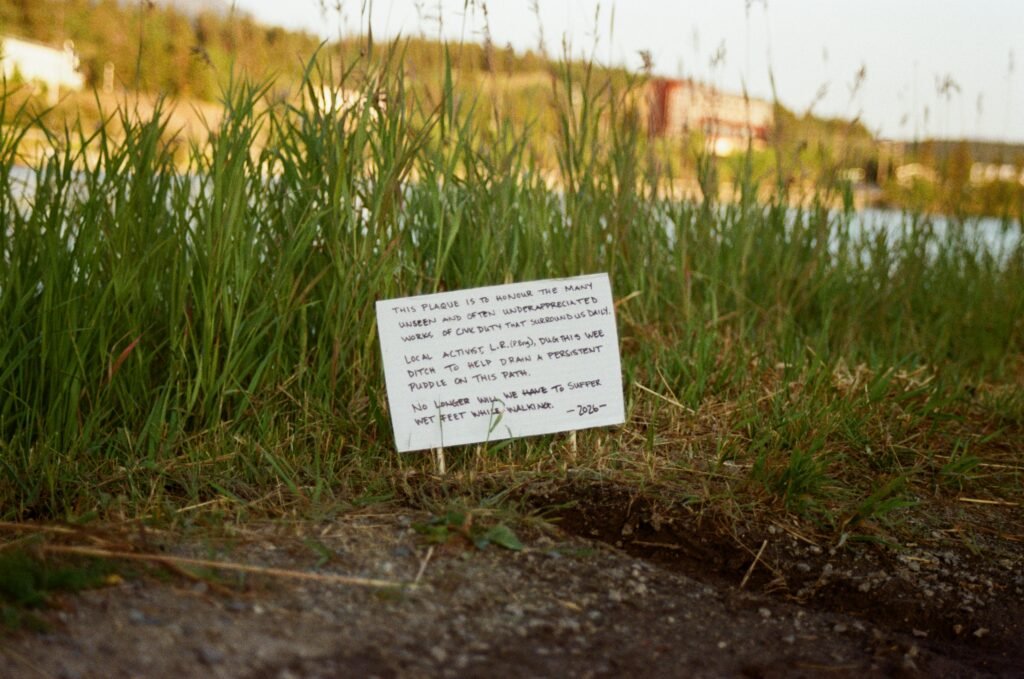 a sign on the ground with a tiny hand-dug ditch. It reads:
This plaque is to honour the many unseen and often underappreciated works of civic duty that surround us daily. Local activist L.R. (P.Eng.) dug this wee ditch to help drain a persistent puddle on this path. No longer will we have to suffer wet feet while walking. -2026-
