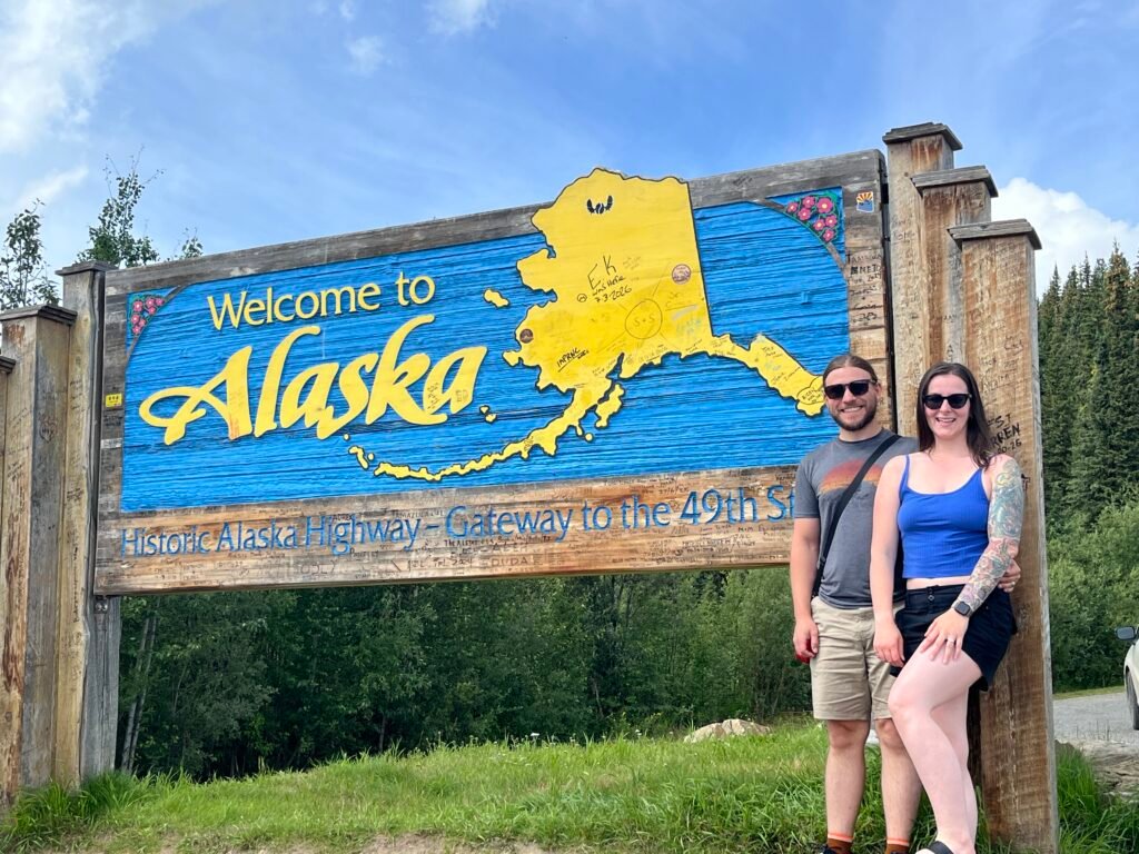two people standing in front of a sign saying "Welcome to Alaska - Historic Alaska Highway"